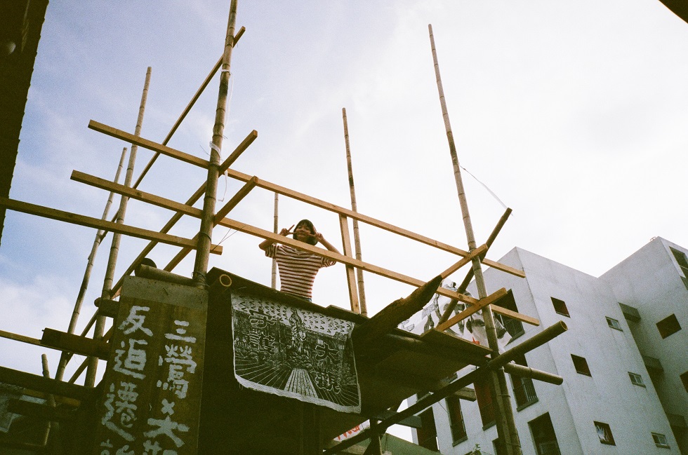 「反迫遷高台」是三鶯部落族人為大觀搭建起的。照片中的慧泉常拉著阿嬤一起參與抗爭，很早就學會了要求警察出示警編、背出逮捕抗爭者所依據的法條，她是自救會其中一位美術股長。