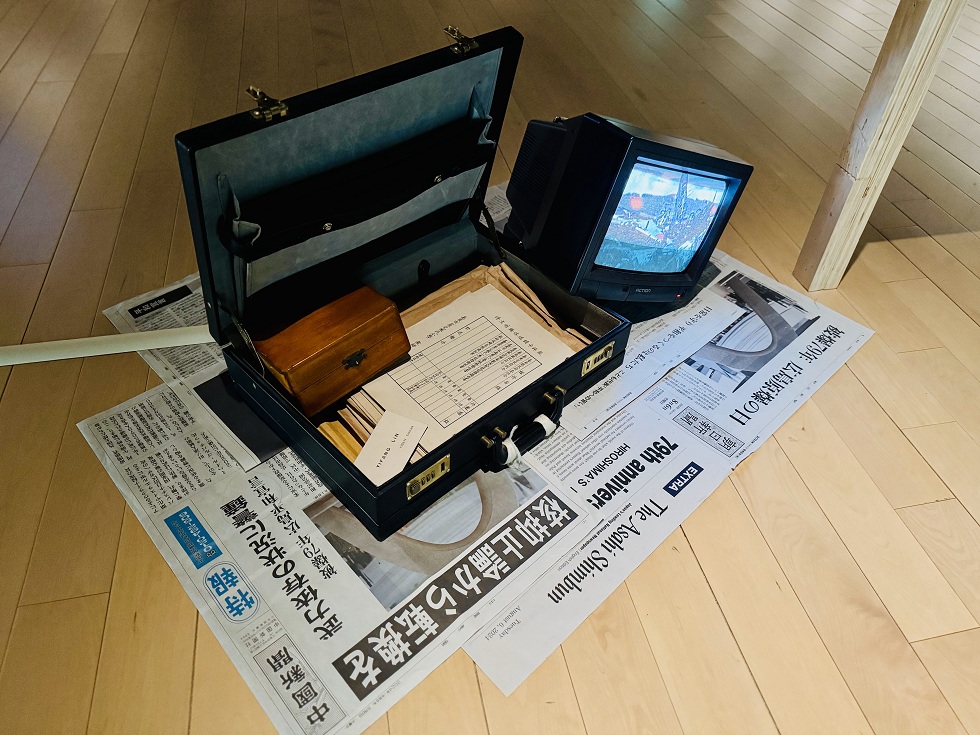 展覽中裝滿文件與一個小木盒的手提箱，底下是關於「79周年原爆紀念」的號外報紙，一旁的電視則播放著零式戰機墜毀的殘骸影像。